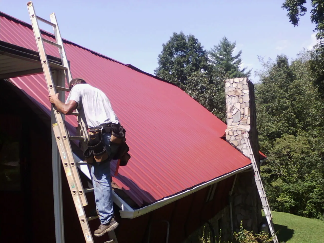 Roofer on a ladder beside a red metal roof and stone chimney — Soffit & Fascia Repair by Roofing Brothers in Coal Township